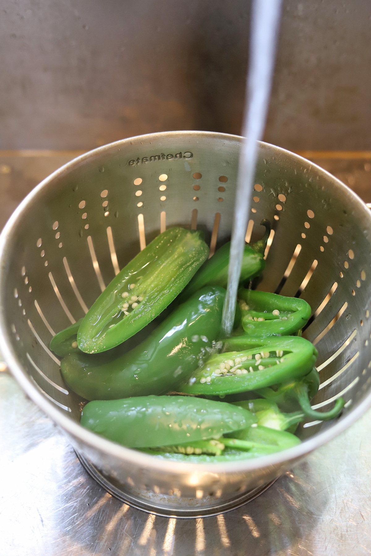 Jalapeno pepper halves in colander with cold water pouring over them.