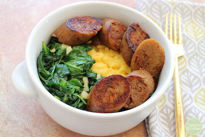 Sliced Field Roast apple sauce sausage with spinach on polenta in bowl.
