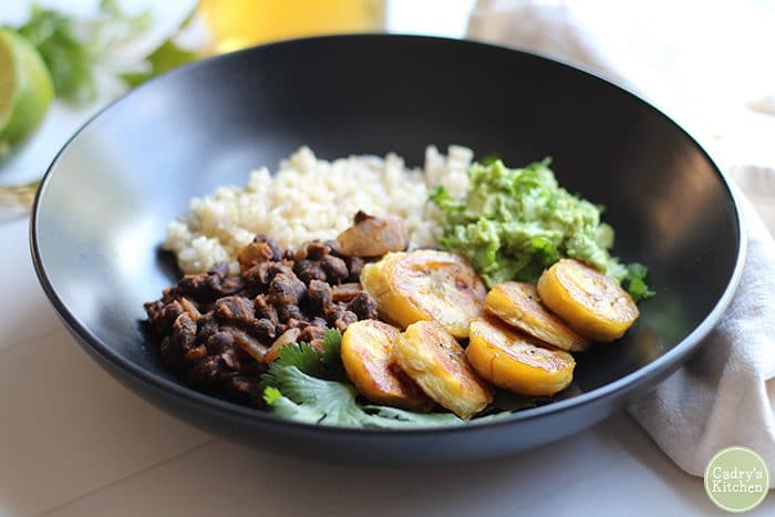 Beans, rice, fried plantains and guacamole in black dish on table.