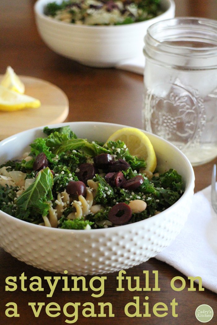Text overlay: Staying full on a vegan diet. Pasta in bowl with sauteed kale, lemon, kalamata olives, and beans.