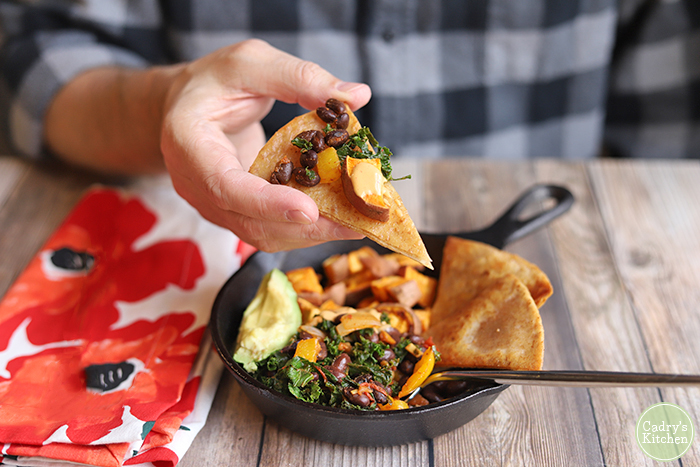 Hand holding fried tortilla wedge topped with sweet potato, kale, and beans.