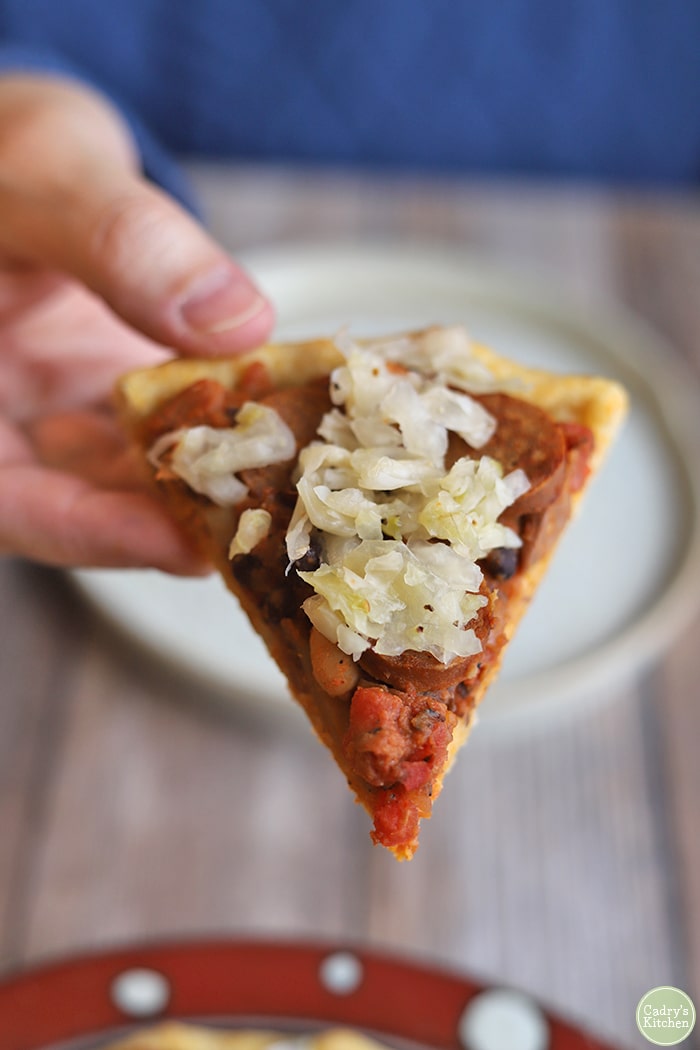 Hand holding slice of vegan chili dog pizza with sauerkraut.