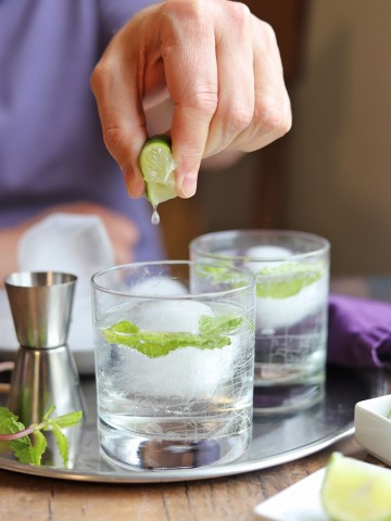 Fingers squeezing lime over gin and tonic with mint.