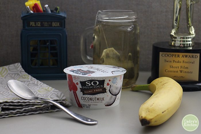 Container of So Delicious coconut milk yogurt and a banana on desk at work.