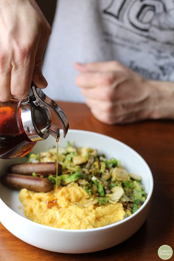Maple syrup being poured on creamy breakfast polenta with vegan sausage & roasted Brussels sprouts.