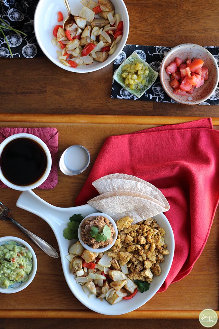 Overhead breakfast potatoes, tomatoes, guacamole, and refried beans on table.