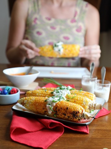 Buffalo corn on the cob with a vegan blue cheese topping on a red napkin. Cadry in background holding ear of corn.