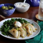 Browned pierogi on a white plate with kale & cashew cream cheese. In the background, a green napkin, water glass, blue bowl, and olives.