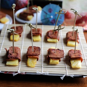 Seitan bacon wrapped pineapple on bamboo covered plate, parasols in background.
