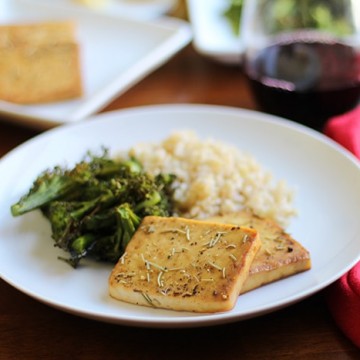 Baked tofu with lemon & rosemary on plate with broccolini & brown rice. Red wine & red napkin on table.