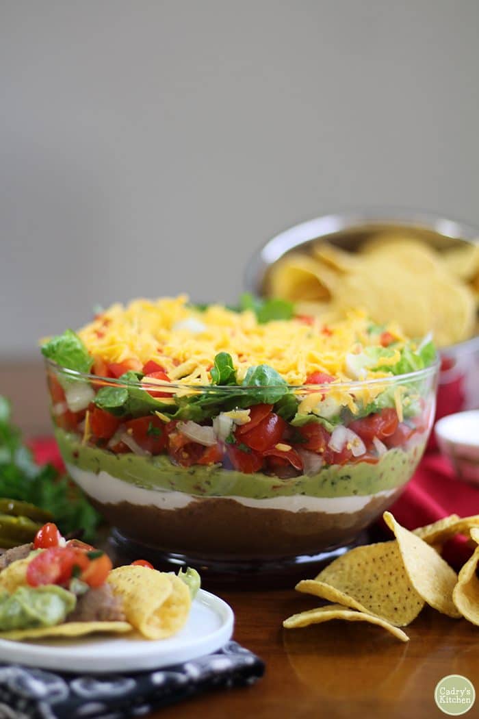 Vegan 7 layer dip in glass bowl with tortilla chips & small plate.