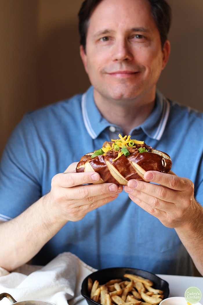 David holding vegan chili dog on a pretzel bun with non-dairy cheese and three bean chili.