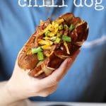 Hand holding vegan chili dog with non-dairy cheese and onions.