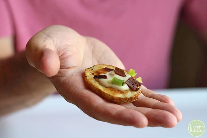 Hand holding loaded potato slice with cashew cream & seitan bacon.