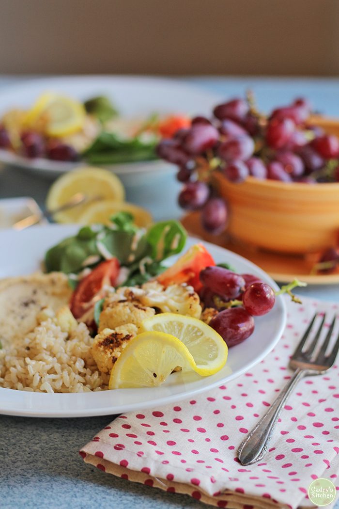 Lemon slice on vegan brown rice bowl with grapes, rice, hummus, and spinach salad.