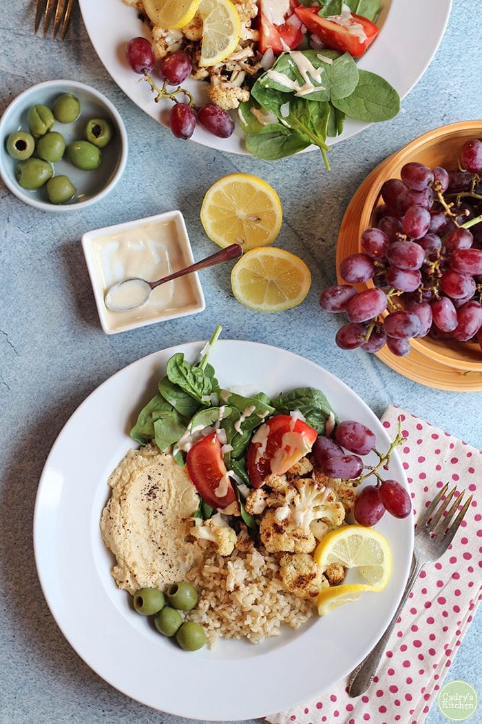 Overhead brown rice bowl with lemons, hummus, tahini sauce, grapes, and Castelvetrano olives. Polka dot napkin & fork next to plate.