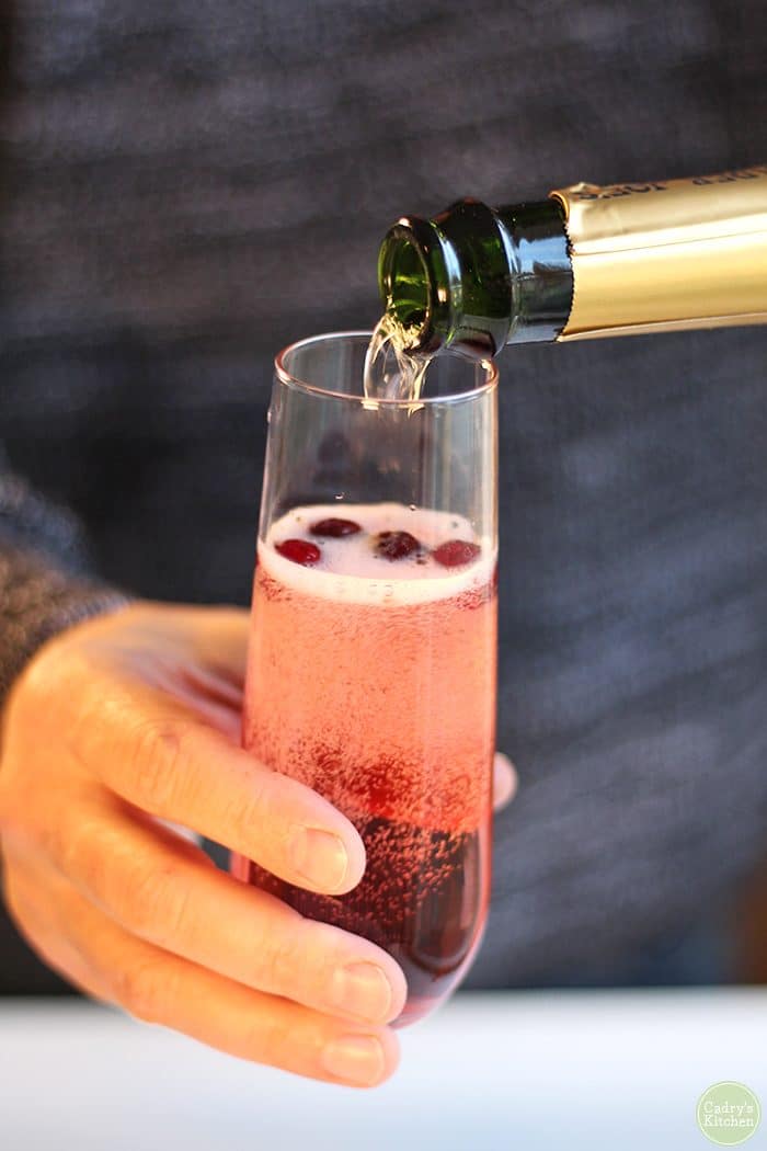 Hand pouring sparkling wine into glass with cranberries and cranberry juice.
