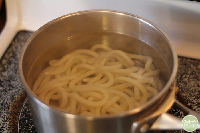 Frozen udon noodles heating in boiling water on stove.