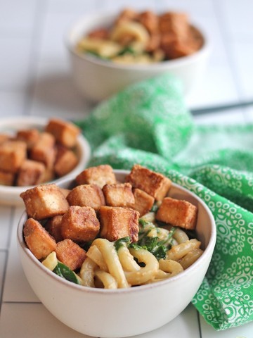 Bowl of peanut butter noodles topped with fried hoisin tofu & sauteed spinach by green napkin.