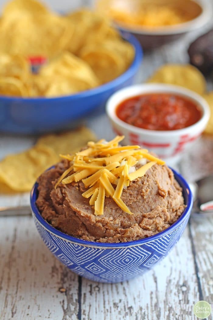 Refried pinto beans covered in non-dairy cheese in blue bowl. Chips and salsa in background.