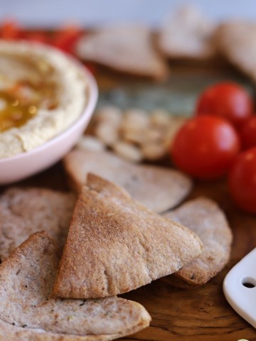 Close-up homemade pita chips on board with hummus, tomatoes, and Marcona almonds.