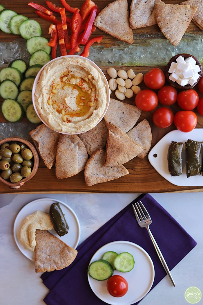 Overhead board with hummus, cucumbers, olives, dolmas, pita chips, and purple napkin.