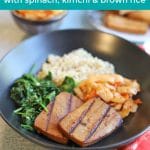 Text: Bulgogi tofu bowls with spinach, kimchi, and brown rice. Bowl with tofu, spinach, kimchi, and rice on table.