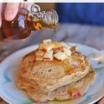 Text overlay: Apple cinnamon pancakes. Vegan. Syrup being poured onto stack of pancakes.