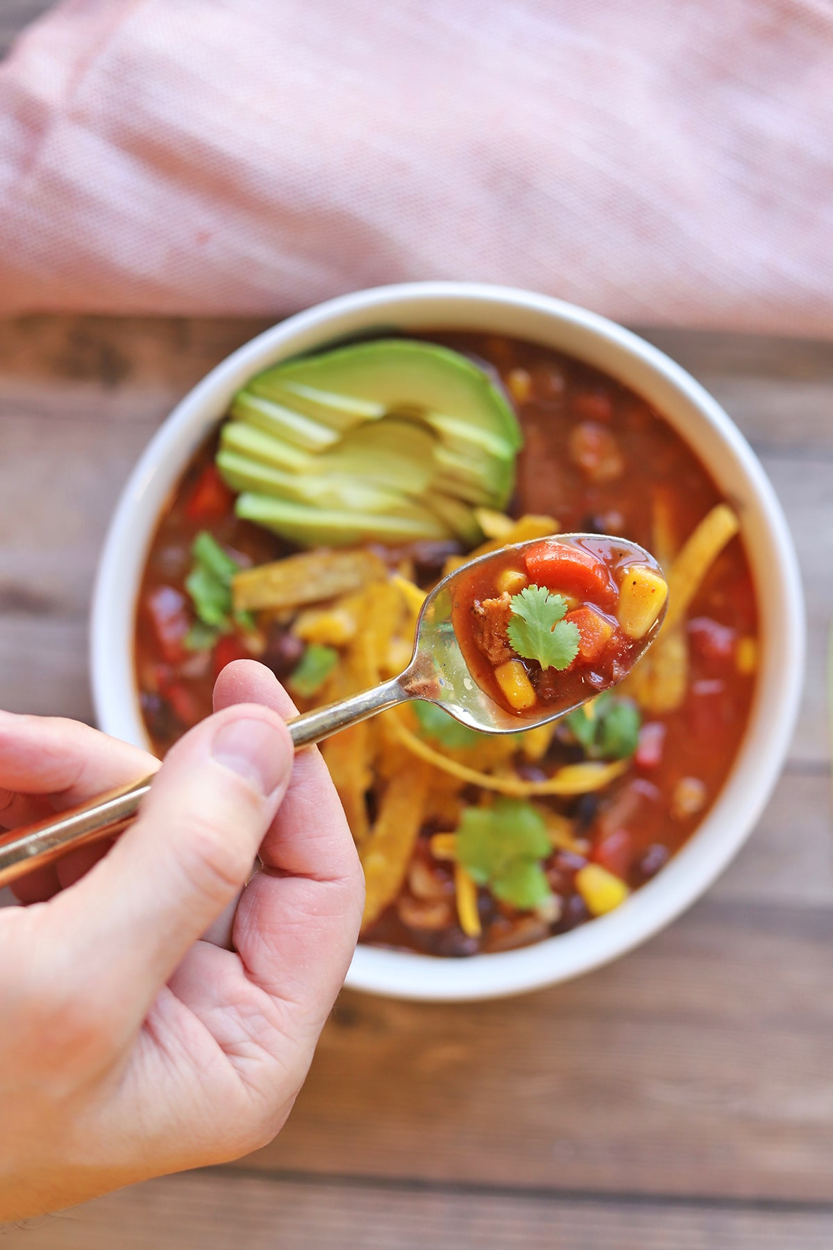 Overhead spoon over bowl of soup with avocado, cilantro, and crispy tortilla strips.