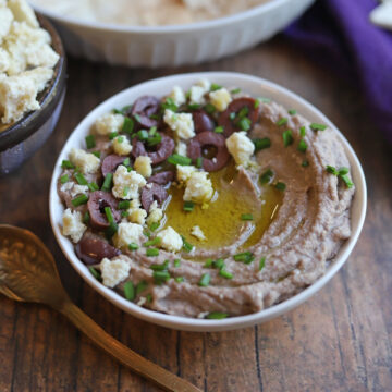 Creamy kalamata olive hummus in bowl, garnished with extra virgin olive oil, vegan feta cheese, and kalamata olives.