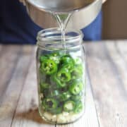 Sugar-free quick pickling brine being poured over sliced jalapenos and garlic in jar.