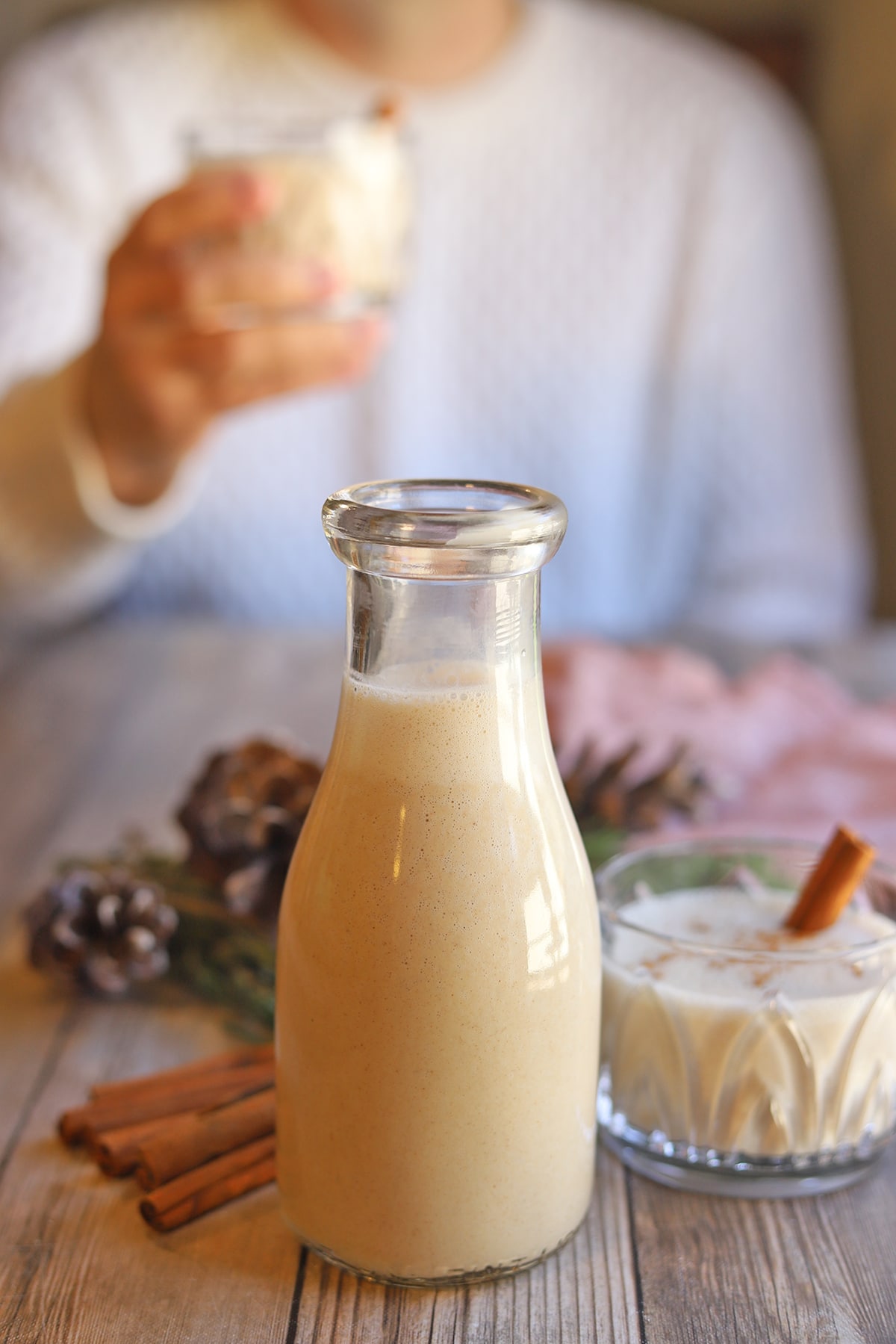 Glass milk bottle on table filled with vegan eggnog. Man drinking glassful of nog in background.