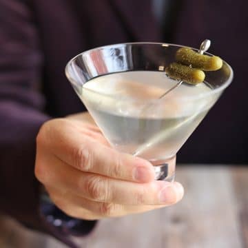 Hand holding martini glass with cornichon garnish.