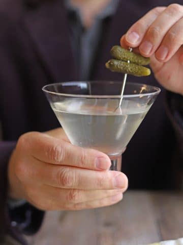 Hand lifting cornichon garnish from martini glass.
