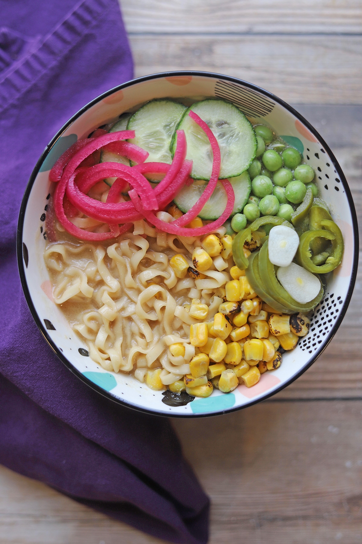 Overhead ramen noodle soup with corn, jalapenos, pickled onions, cucumbers, and peas.