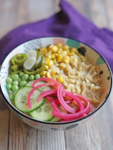 Bowl of vegan ramen noodles with corn, jalapenos, peas, cucumbers, and pickled onions.