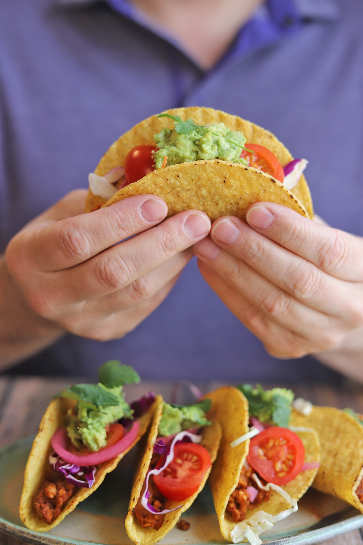 Hand holding hard shell taco over platter of tacos.