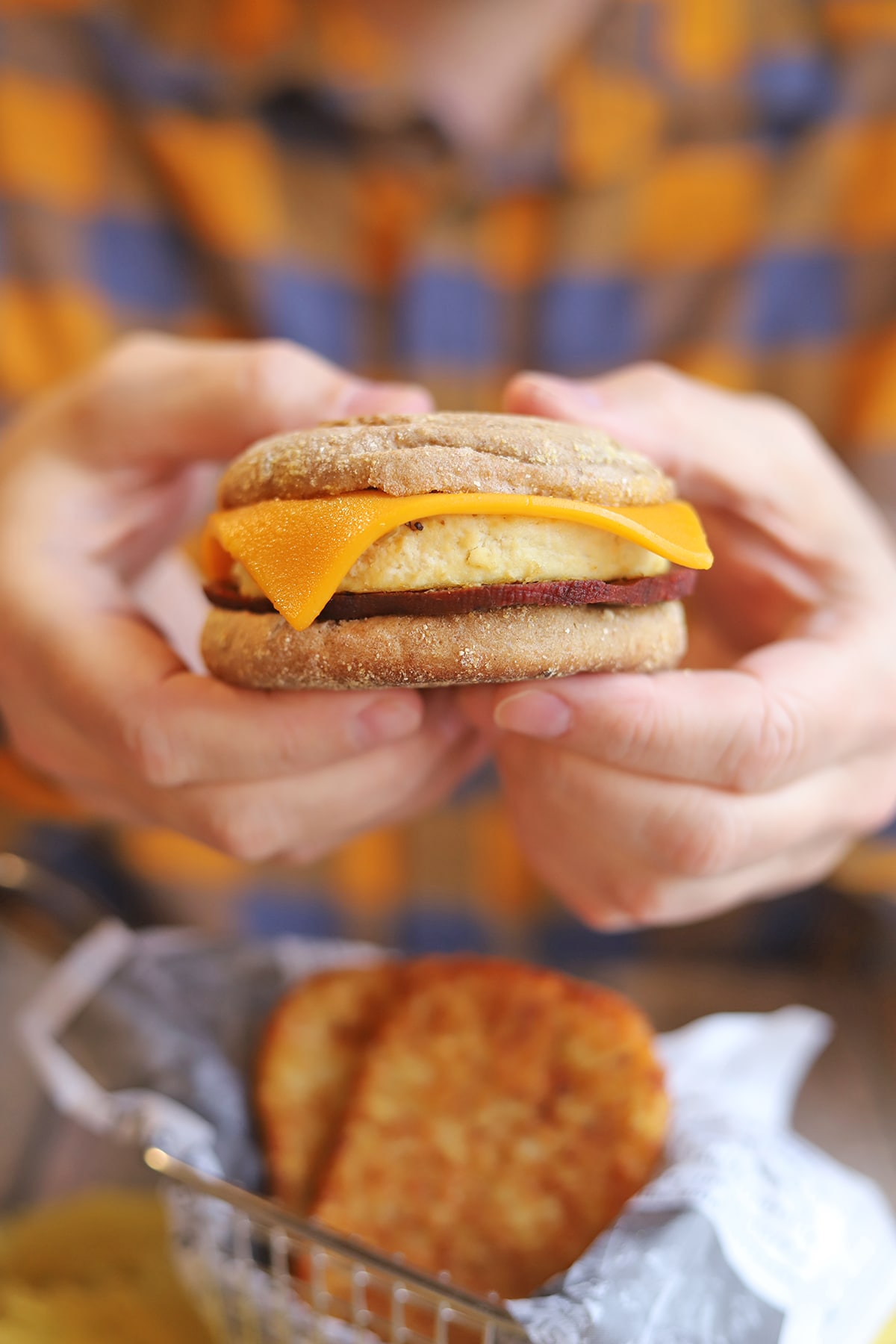 Hands holding English muffin breakfast sandwich by hashbrown patties.