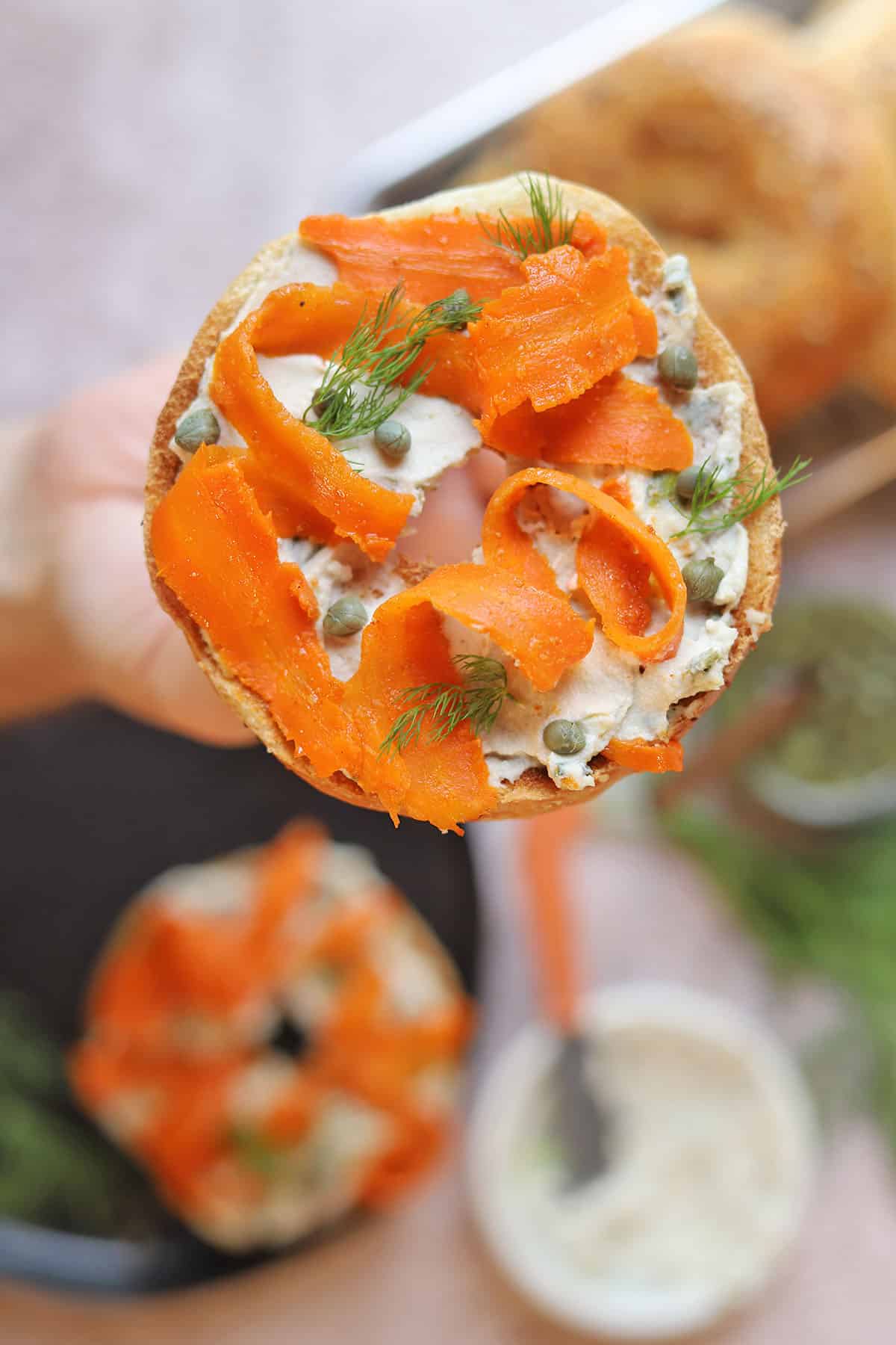 Hand holding bagel with vegan lox, cream cheese, and dill.