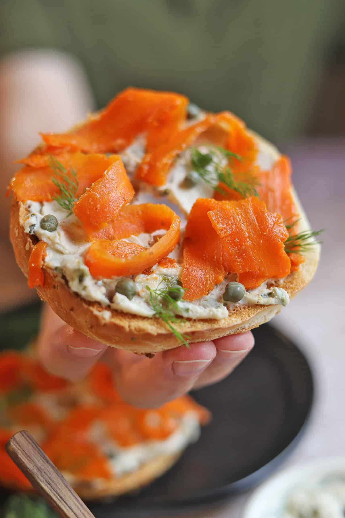 Hand holding bagel with vegan lox, non-dairy cream cheese, capers, and dill.