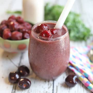 Cherry smoothie in glass by frozen cherries, spoon, and colorful napkin.