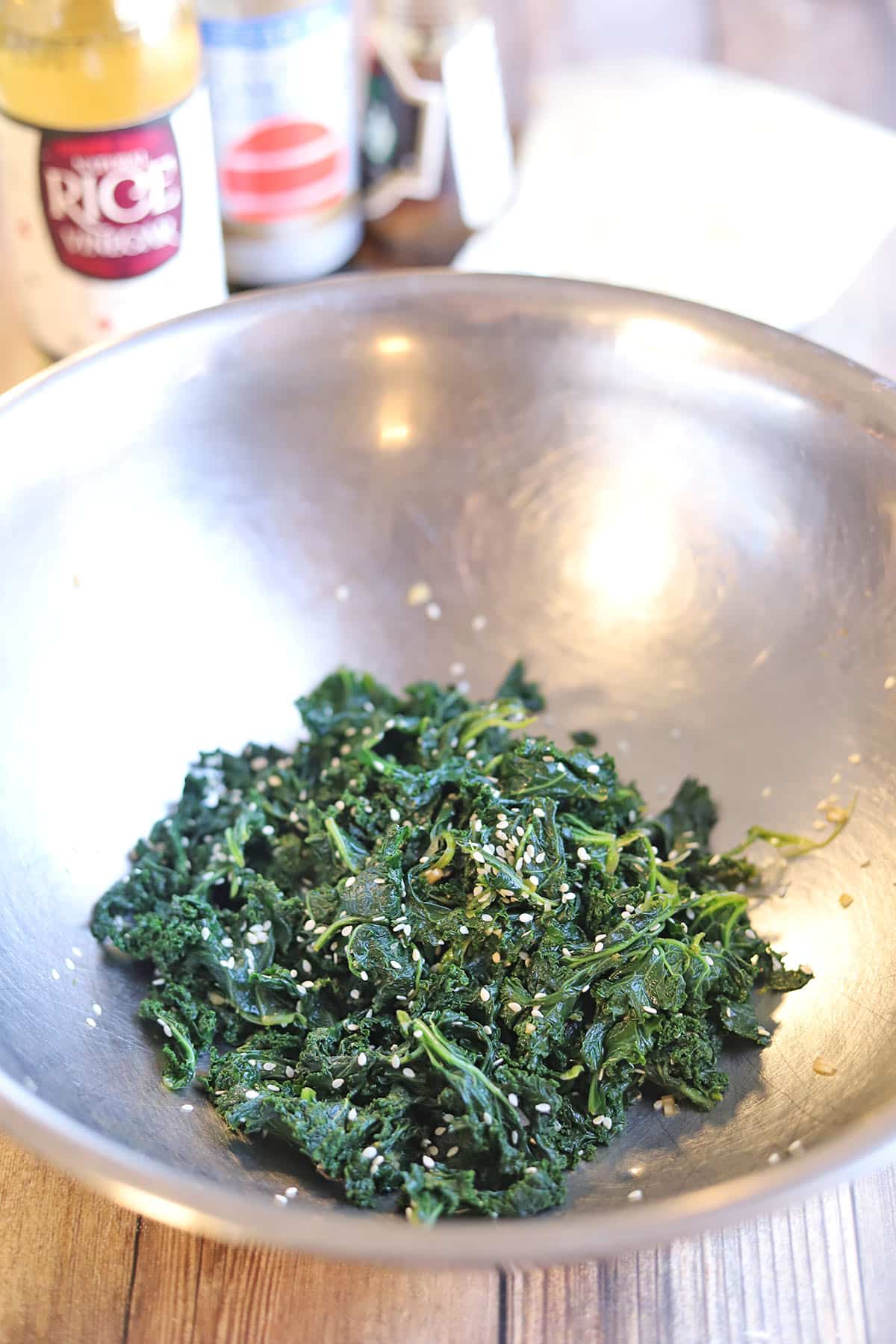 Steamed kale in mixing bowl with sesame oil, rice vinegar, tamari, and sesame seeds.
