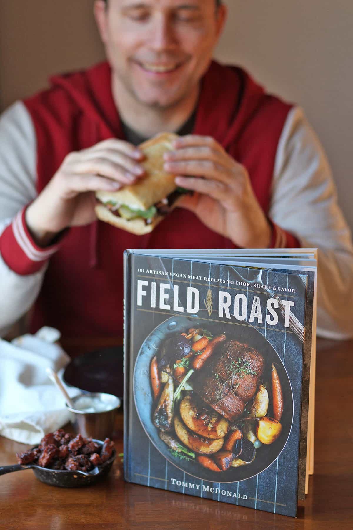 Field Roast cookbook on table. Man smiles holding sandwich in background.