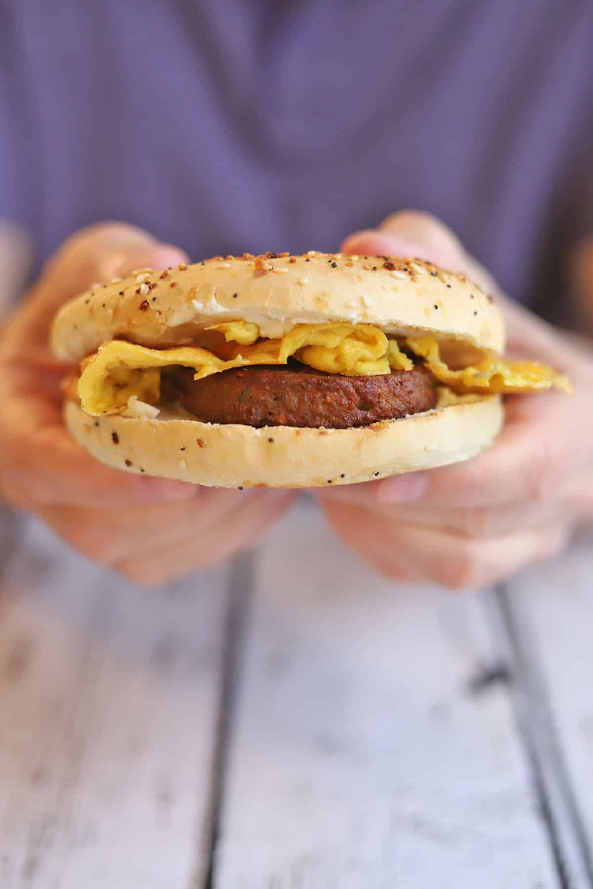 Hand holding bagel sandwich with Just Egg, vegan sausage, and vegan cream cheese.