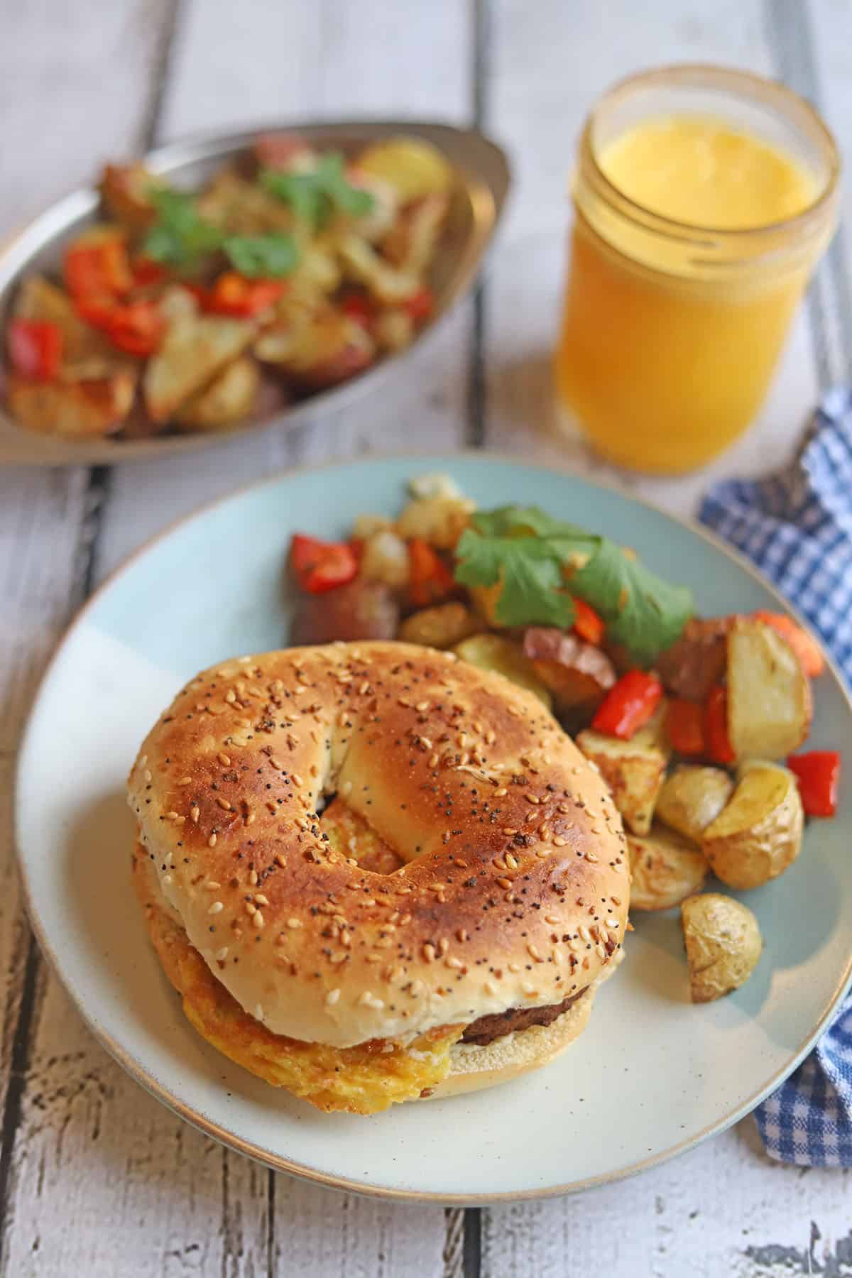 Bagel sandwich on plate with potato hash. On the side, a glass of orange juice.