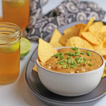 Bowl of vegan chili cheese dip on table with beer and tortilla chips.