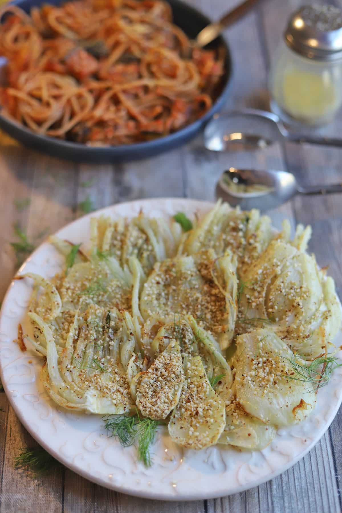 Plate with slices of roasted fennel in front of pasta bowl and tongs.
