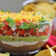 Text overlay: Vegan 7-layer dip. Close-up salsa, guacamole, sour cream, and refried beans in glass bowl.