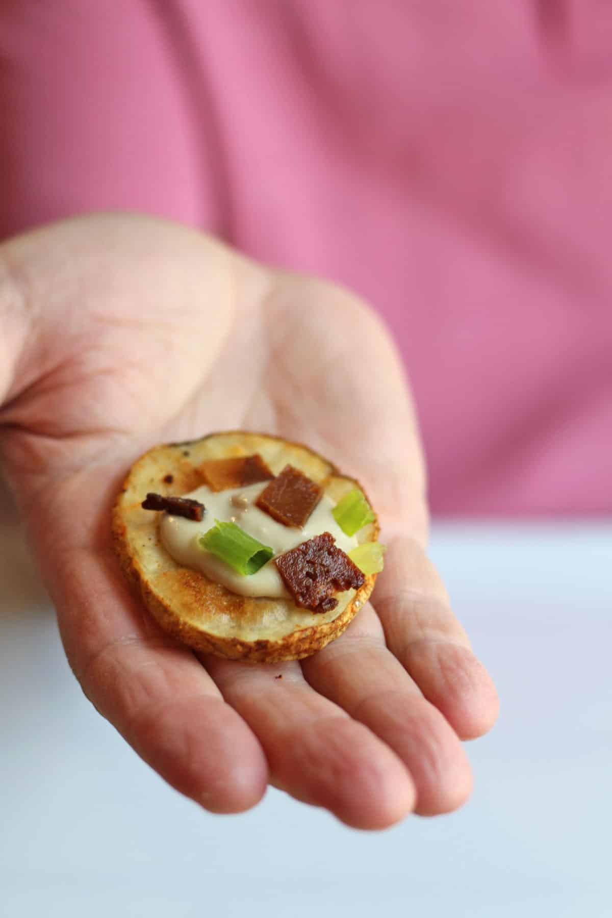 Hand holding air fried potato slice with cashew cream, seitan bacon, and green onion.