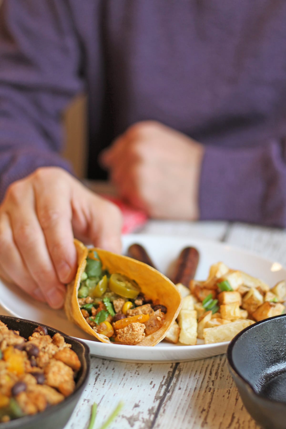 Hand holding vegan tofu taco on plate with seitan sausage links and potatoes.